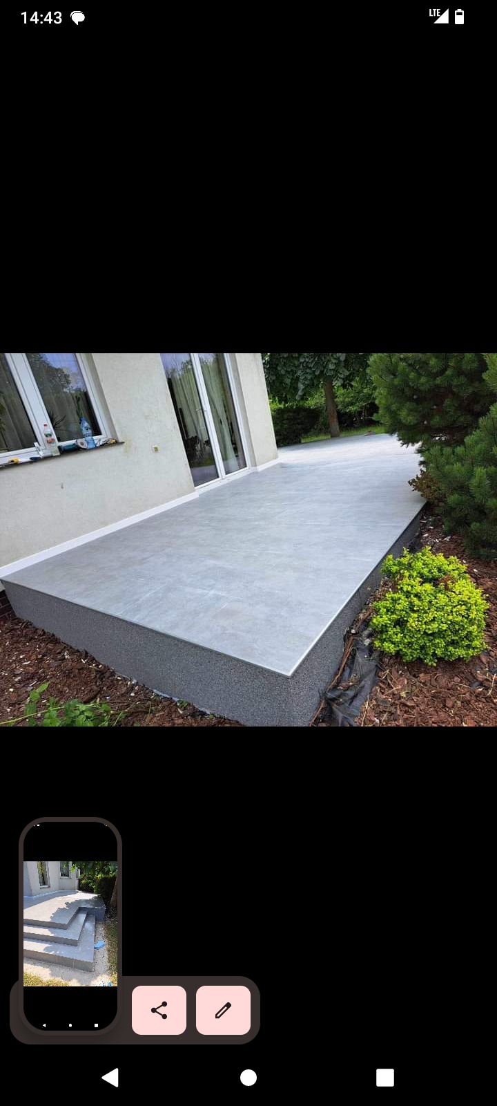 Wykończony taras z szarą, gładką powierzchnią, obłożony drobnym kamieniem, przylegający do białej ściany budynku z oknami i drzwiami tarasowymi, otoczony zielenią.