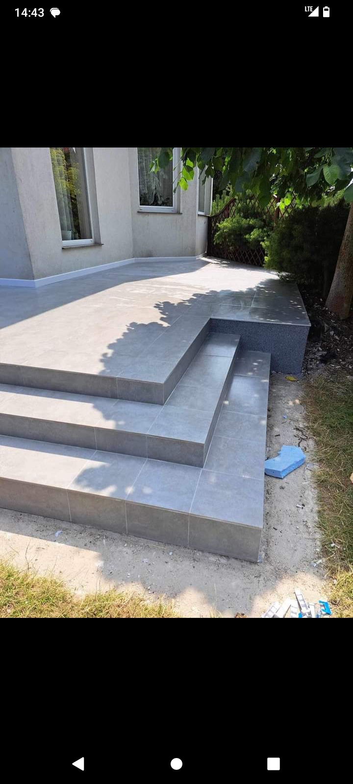 Wykończony taras z szarych płytek ceramicznych, prowadzący schodami do ogrodu, z widocznym fragmentem elewacji budynku i zielenią.