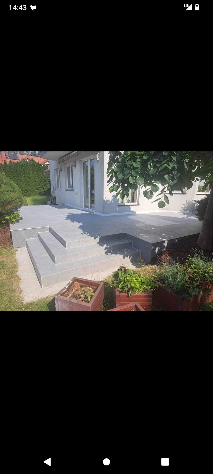 Nowoczesny taras z szarych płytek, schody z trzech stopni, drewniane donice z zielenią, budynek w tle, częściowo zacieniony przez drzewo.