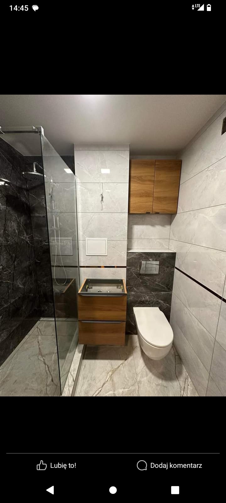 Nowoczesna łazienka w trakcie montażu: kabina prysznicowa ze szklaną ścianą, podwieszana toaleta i drewniana szafka pod umywalkę bez blatu, marmurowe płytki na podłodze i ścianach.