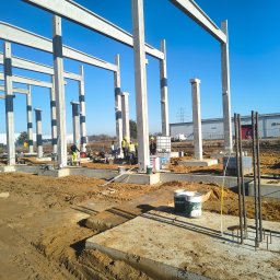 Konstrukcja szkieletowa budynku w trakcie budowy, widoczne betonowe słupy i belki, zbrojenia, pracownicy w kaskach, teren budowy w słoneczny dzień.