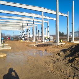 Konstrukcja szkieletowa budynku w trakcie budowy, widoczne betonowe słupy i belki, pracownicy w tle, teren budowy z błotem i materiałami budowlanymi.