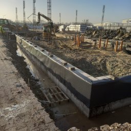 Izolacja fundamentów z folią kubełkową, zbrojenie pionowe, wykop, maszyny budowlane w tle, słoneczny dzień. Widok na narożnik fundamentu.