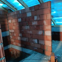 Wznoszenie ściany z czerwonej cegły dziurawki na budowie domu. Widoczne drewniane elementy konstrukcji dachu i niebieska folia. Częściowo widoczne schody i narzędzia.