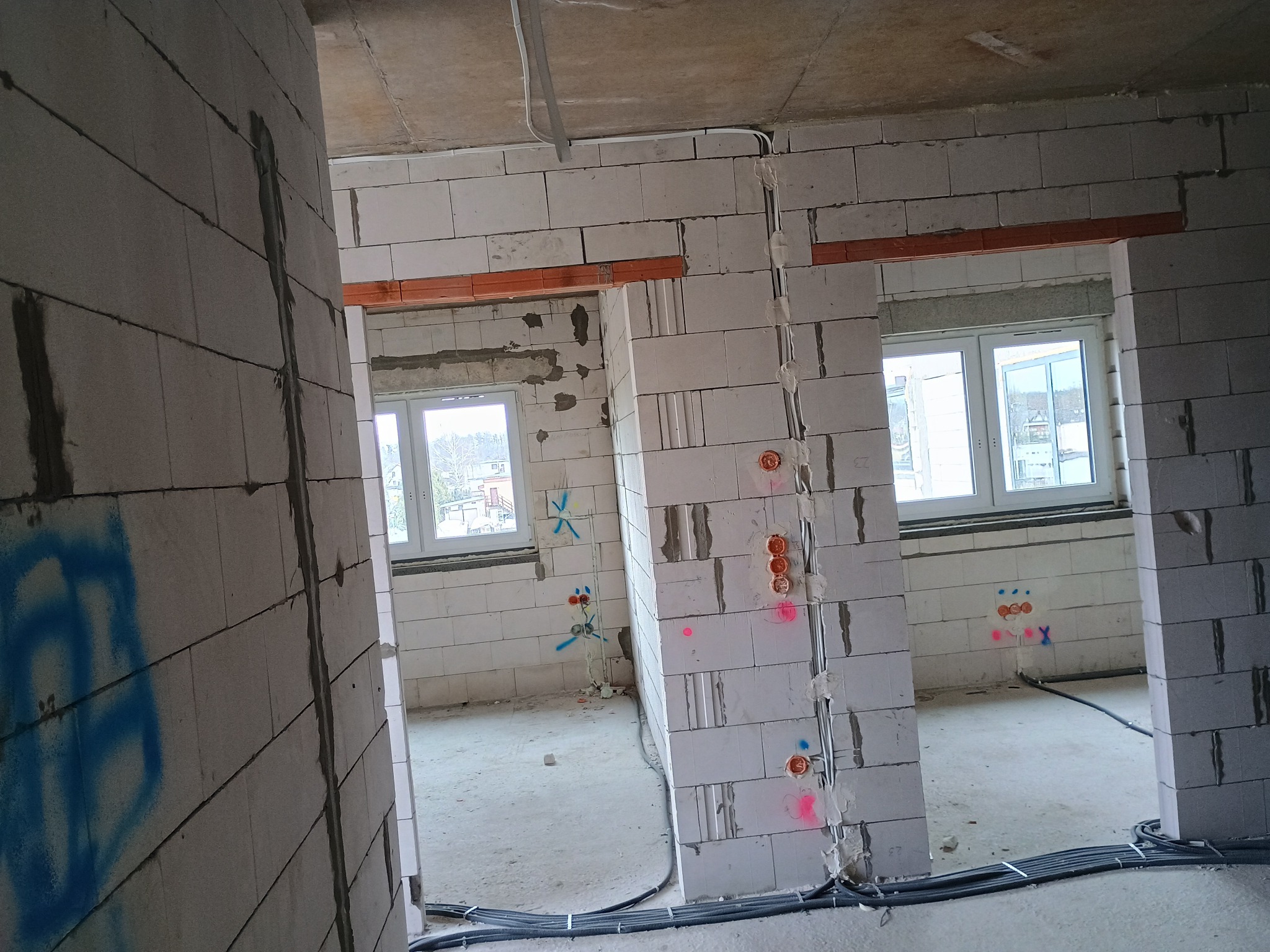 Wnętrze w trakcie budowy: ściany z białych bloczków, widoczne okna, instalacje elektryczne z puszkami i kable na podłodze. Ślady po oznaczeniach na ścianach.