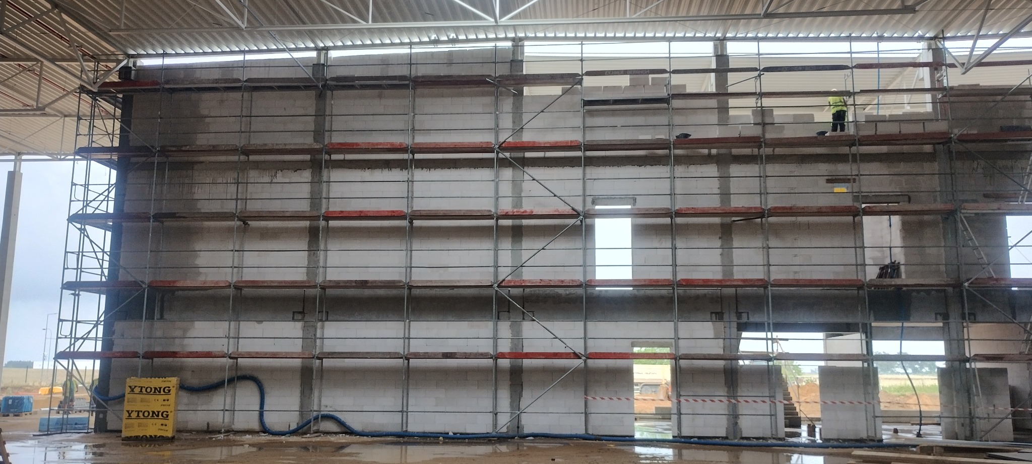 Wznoszenie ściany z bloczków Ytong na rusztowaniu wewnątrz hali. Widoczny pracownik w kamizelce. Na podłodze woda i porozrzucane materiały budowlane.