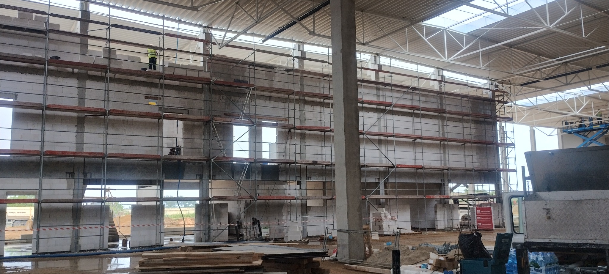 Wnętrze hali w trakcie budowy z białych bloczków, z rusztowaniami i pracownikiem w kamizelce na górze. Widoczna konstrukcja dachu i częściowo sprzęt budowlany.