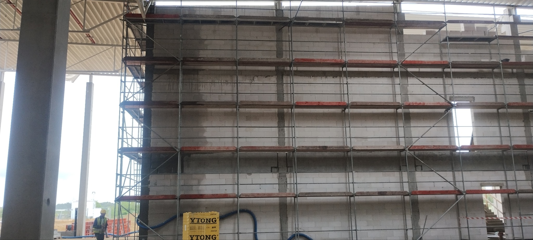 Wznoszenie ściany z białych bloczków Ytong na rusztowaniu. Widoczny pracownik w kasku i kamizelce. Surowy stan budynku, betonowe słupy, konstrukcja dachu.