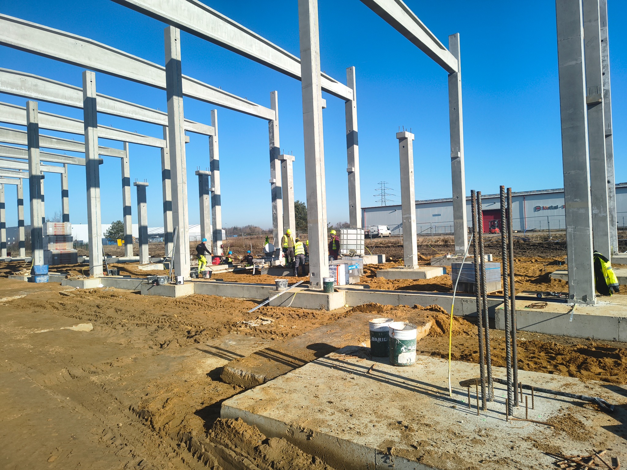 Konstrukcja szkieletowa budynku w trakcie budowy, widoczne betonowe słupy i belki, zbrojenia, pracownicy w kaskach, teren budowy w słoneczny dzień.