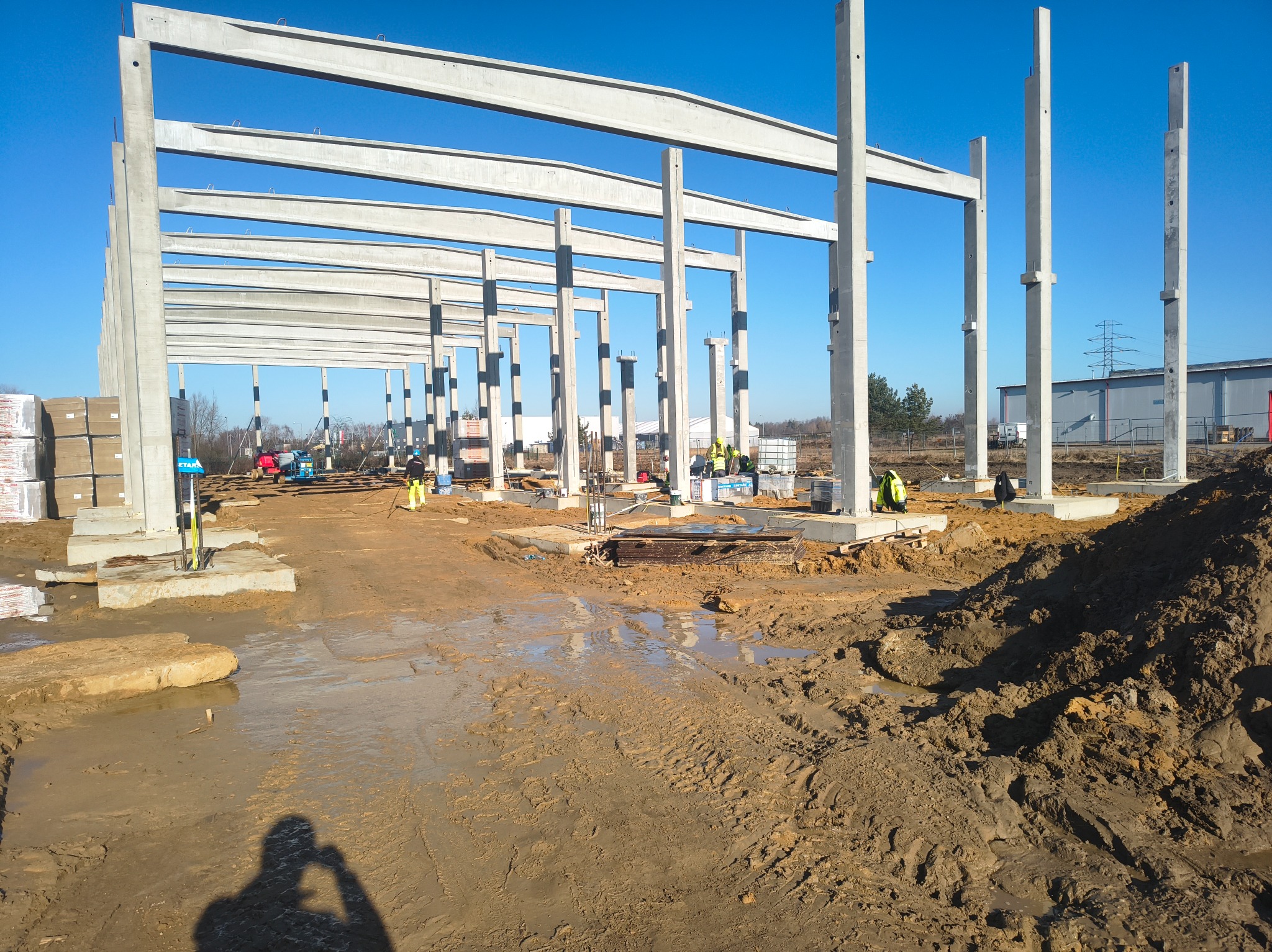 Konstrukcja szkieletowa budynku w trakcie budowy, widoczne betonowe słupy i belki, pracownicy w tle, teren budowy z błotem i materiałami budowlanymi.