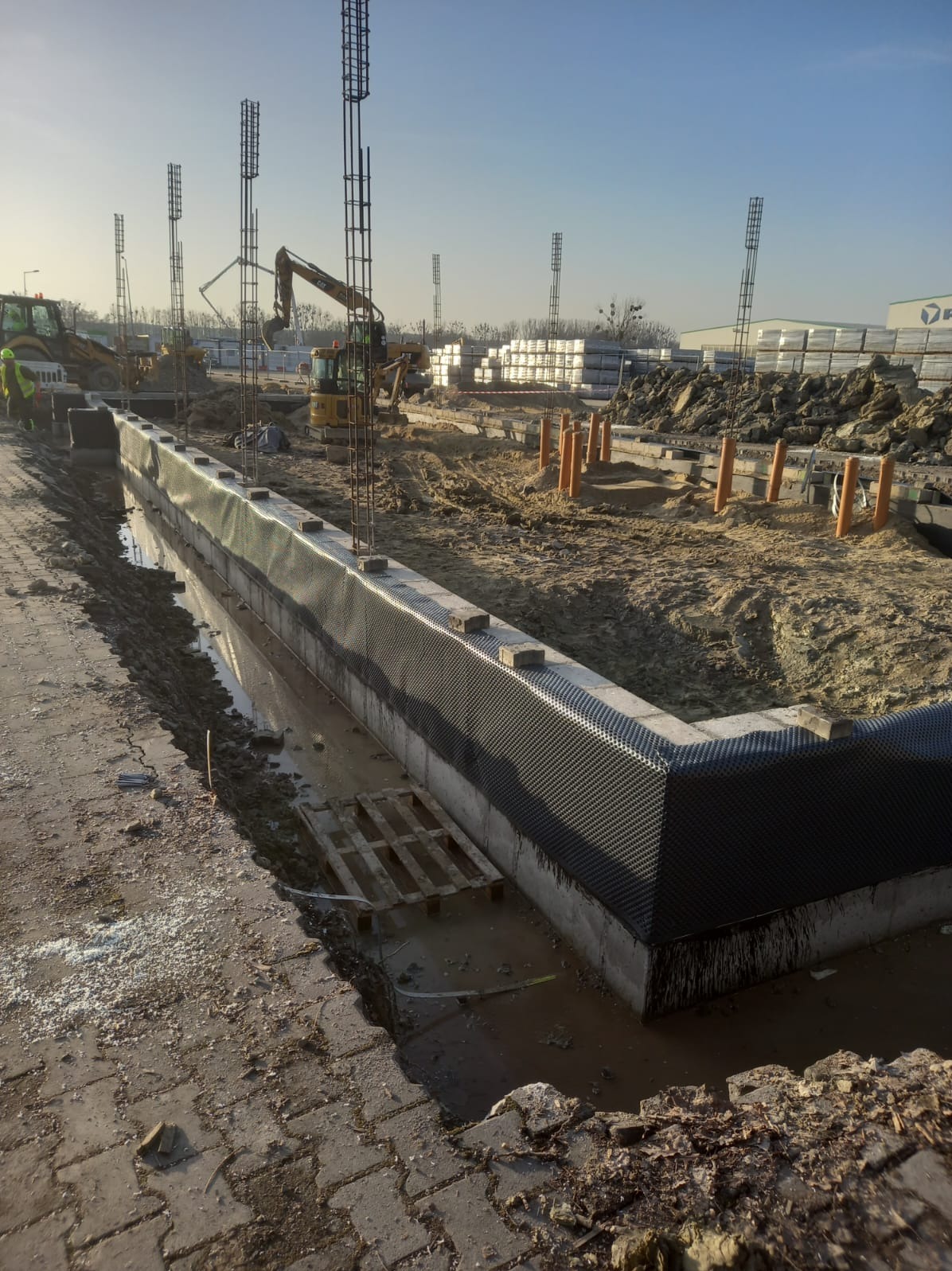 Izolacja fundamentów z folią kubełkową, zbrojenie pionowe, wykop, maszyny budowlane w tle, słoneczny dzień. Widok na narożnik fundamentu.