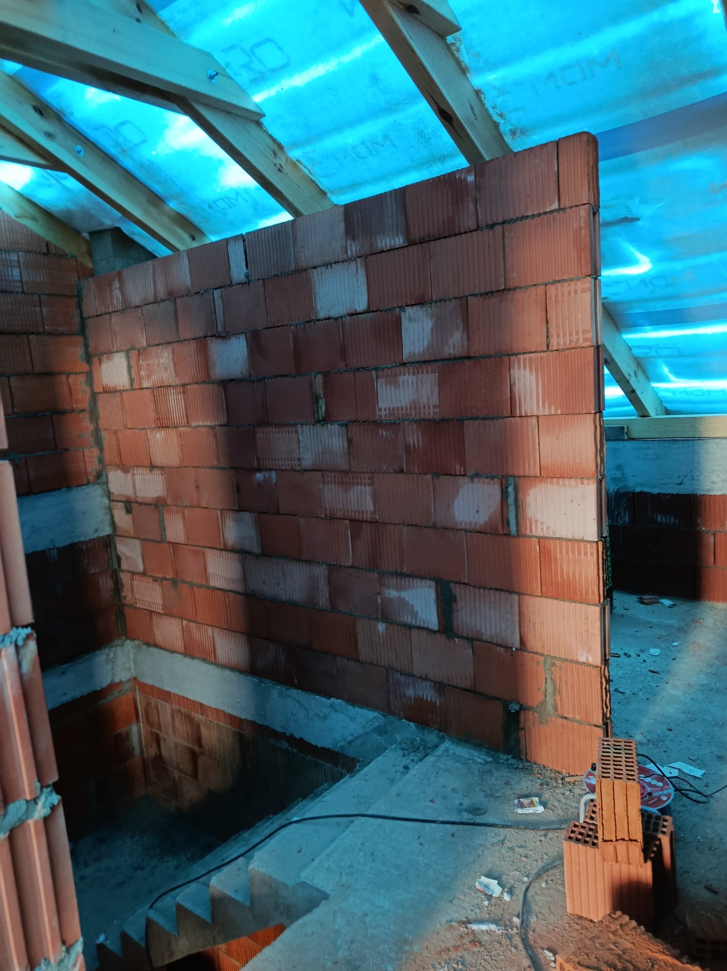 Wznoszenie ściany z czerwonej cegły dziurawki na budowie domu. Widoczne drewniane elementy konstrukcji dachu i niebieska folia. Częściowo widoczne schody i narzędzia.