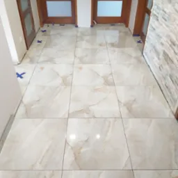 Ułożone jasne płytki podłogowe z marmurkowym wzorem, widoczne niebieskie krzyżyki dystansowe i kliny poziomujące, fragment ściany wykończonej kamieniem dekoracyjnym oraz drewniane ościeżnice...