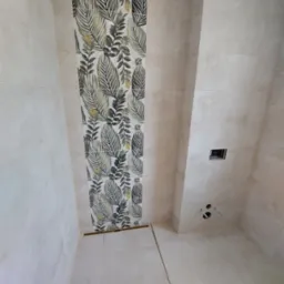 Wykończona łazienka z beżowymi płytkami i dekoracyjnym pasem płytek z motywem liści, widoczny odpływ liniowy w podłodze i przygotowane wyprowadzenia instalacji wodnej.