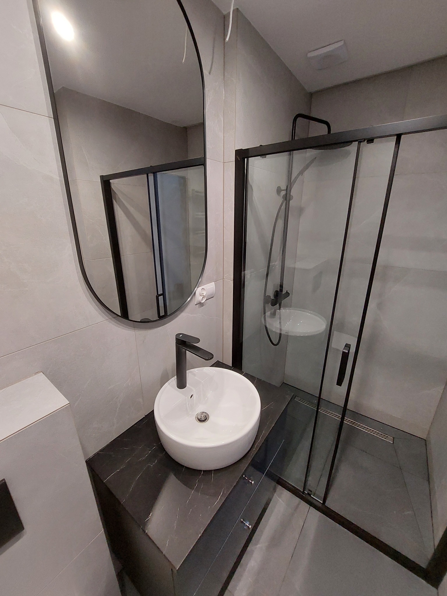 Nowoczesna łazienka z owalnym lustrem, białą umywalką nablatową na czarnym blacie imitującym marmur, kabiną prysznicową typu walk-in z czarnymi profilami i szarymi płytkami.