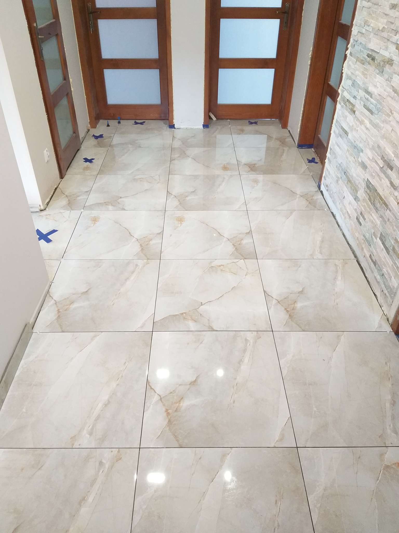 Ułożone jasne płytki podłogowe z marmurkowym wzorem, widoczne niebieskie krzyżyki dystansowe i kliny poziomujące, fragment ściany wykończonej kamieniem dekoracyjnym oraz drewniane ościeżnice...