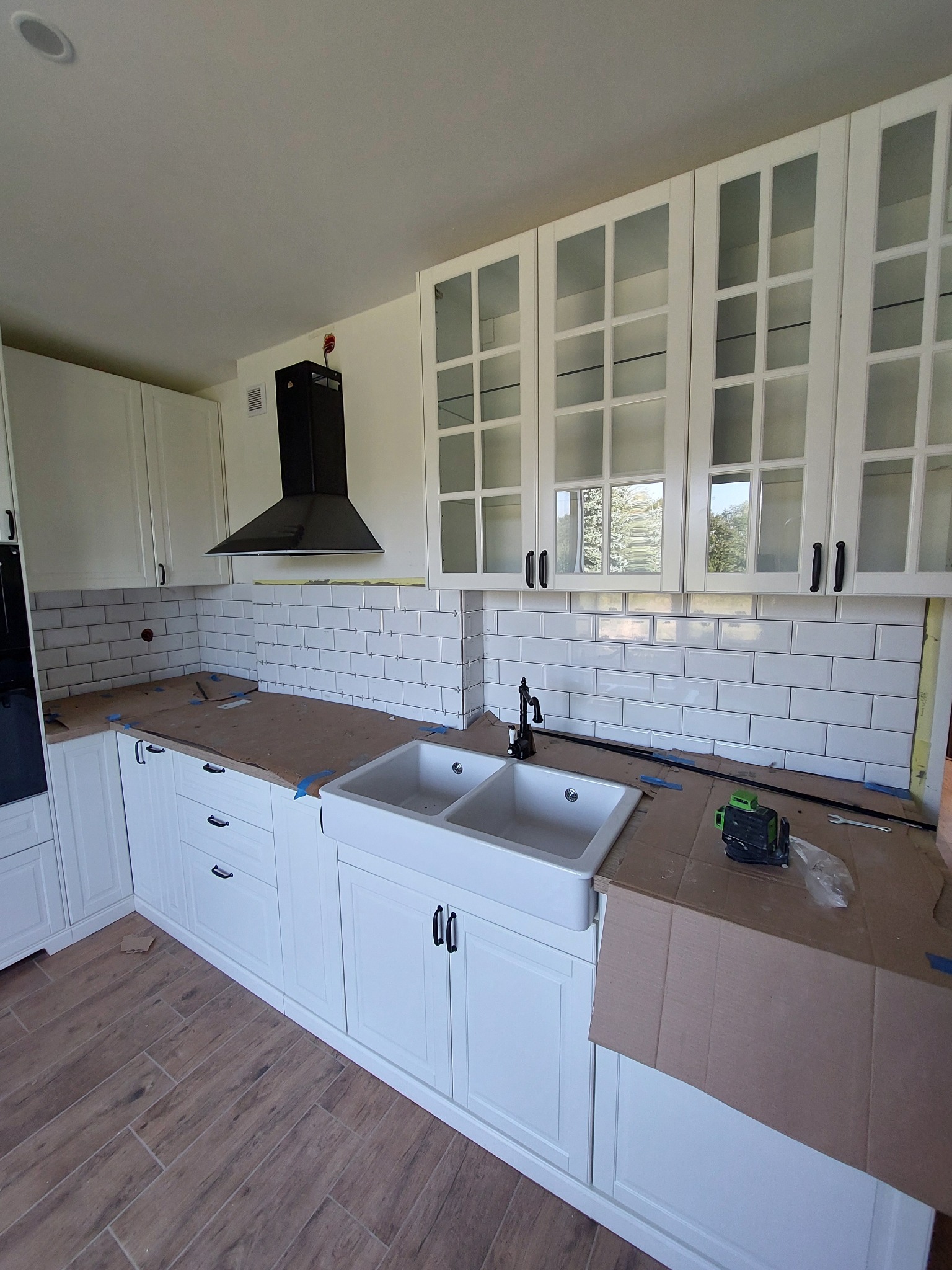 Montaż białych płytek metra na ścianie nad blatem w kuchni z białymi szafkami i czarnym okapem, widoczne narzędzia i zabezpieczenia blatu.