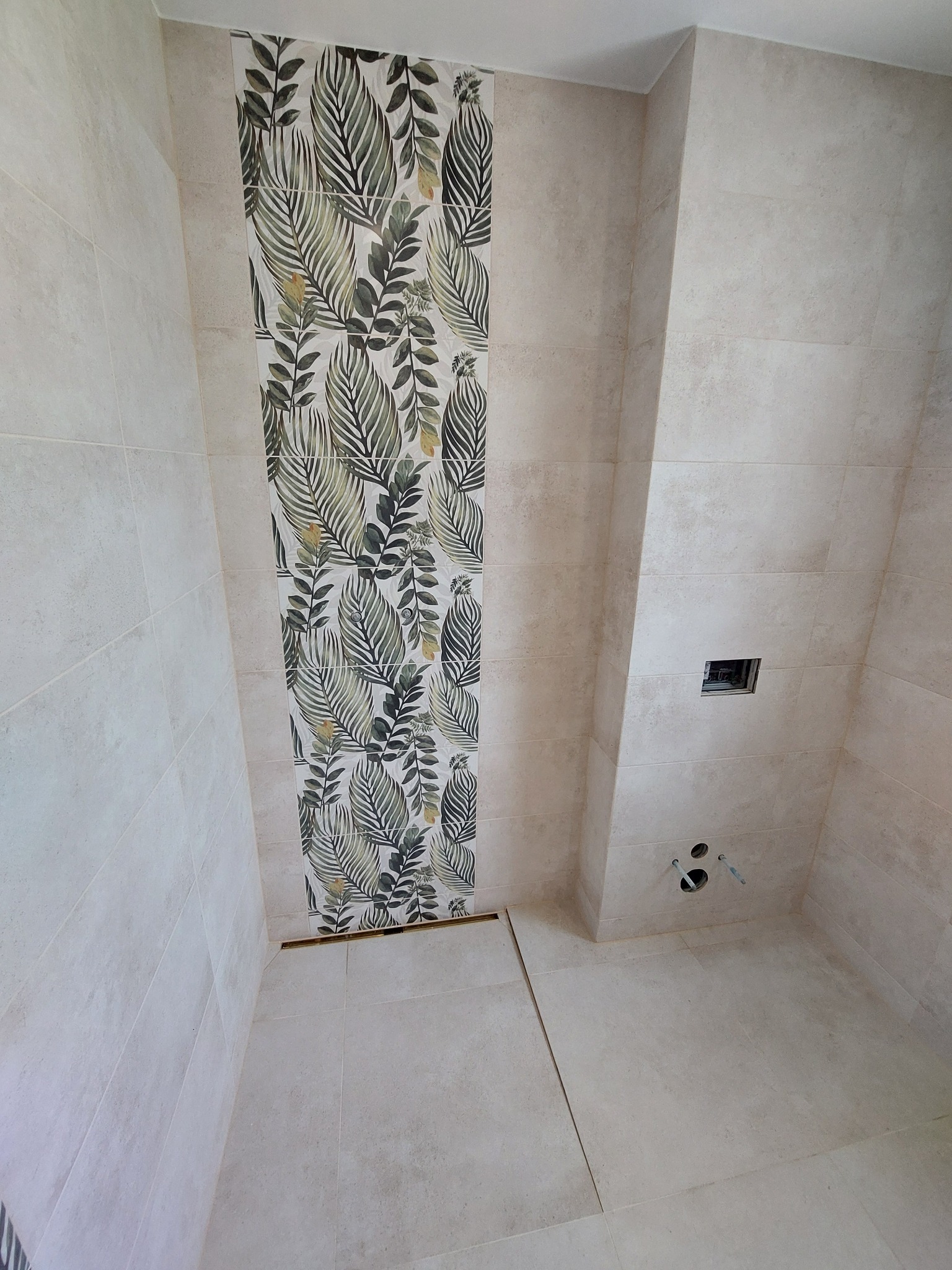 Wykończona łazienka z beżowymi płytkami i dekoracyjnym pasem płytek z motywem liści, widoczny odpływ liniowy w podłodze i przygotowane wyprowadzenia instalacji wodnej.