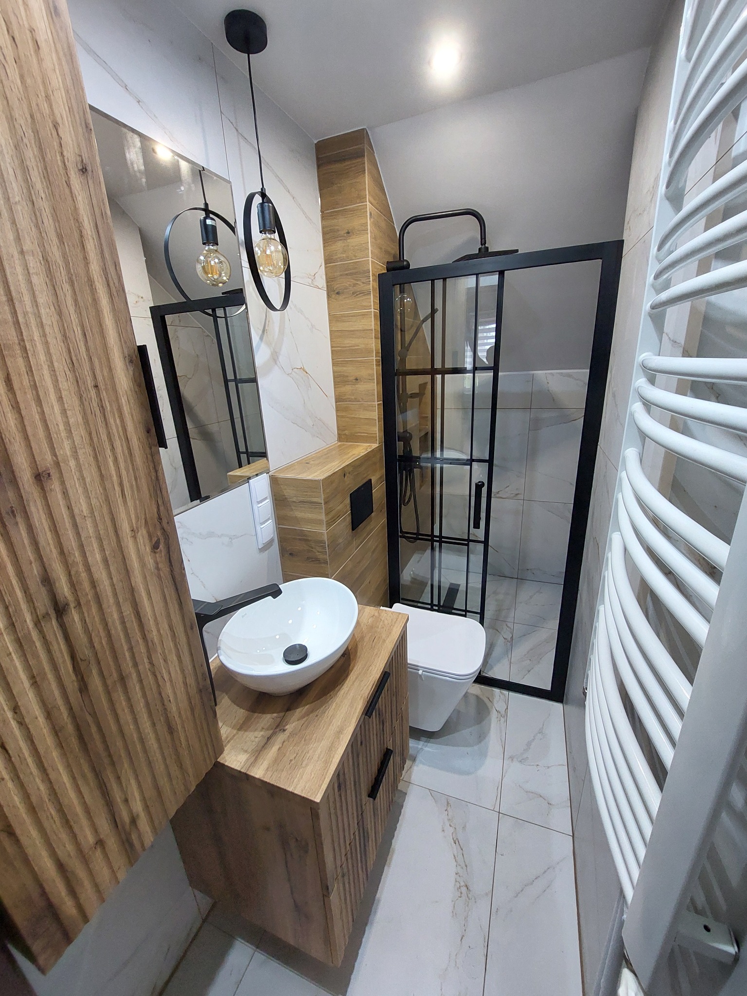 Nowoczesna łazienka z drewnianymi akcentami: umywalka nablatowa na szafce z frezowanymi frontami, kabina prysznicowa typu walk-in z czarnymi profilami, wisząca toaleta, grzejnik drabinkowy...