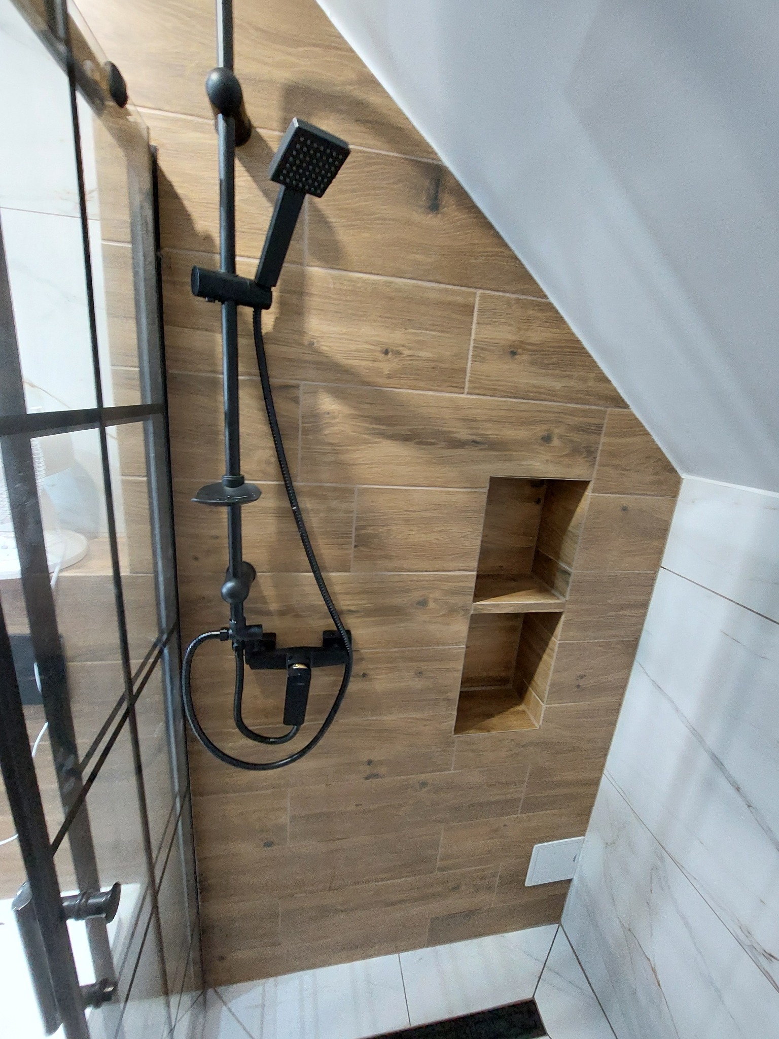 Kabina prysznicowa z czarną armaturą i półkami w ścianie wyłożonej płytkami imitującymi drewno, widok na skośny sufit.