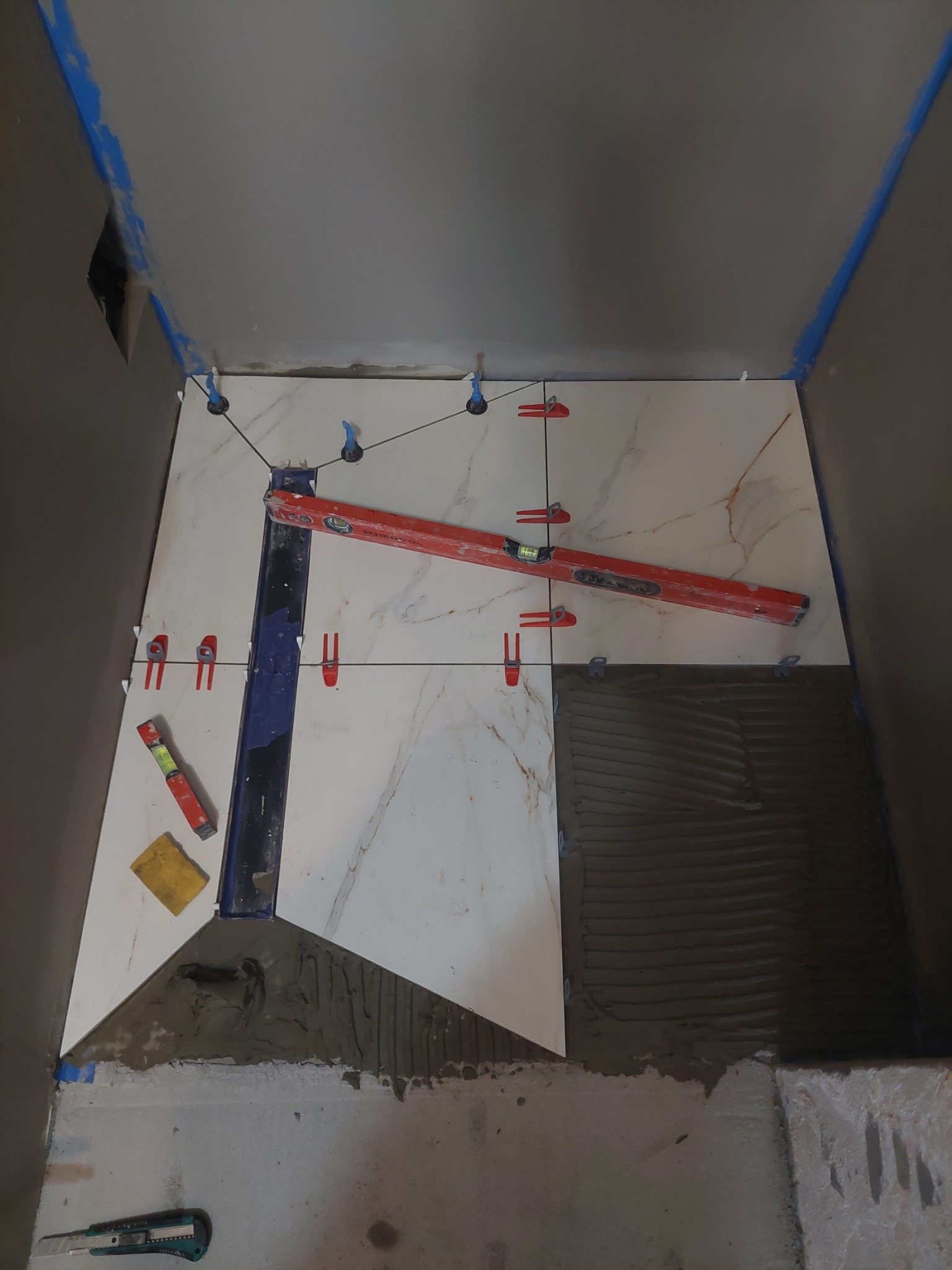 Proces układania płytek imitujących marmur w łazience, widoczne poziomice, szpachelka, nożyk i system poziomowania płytek.