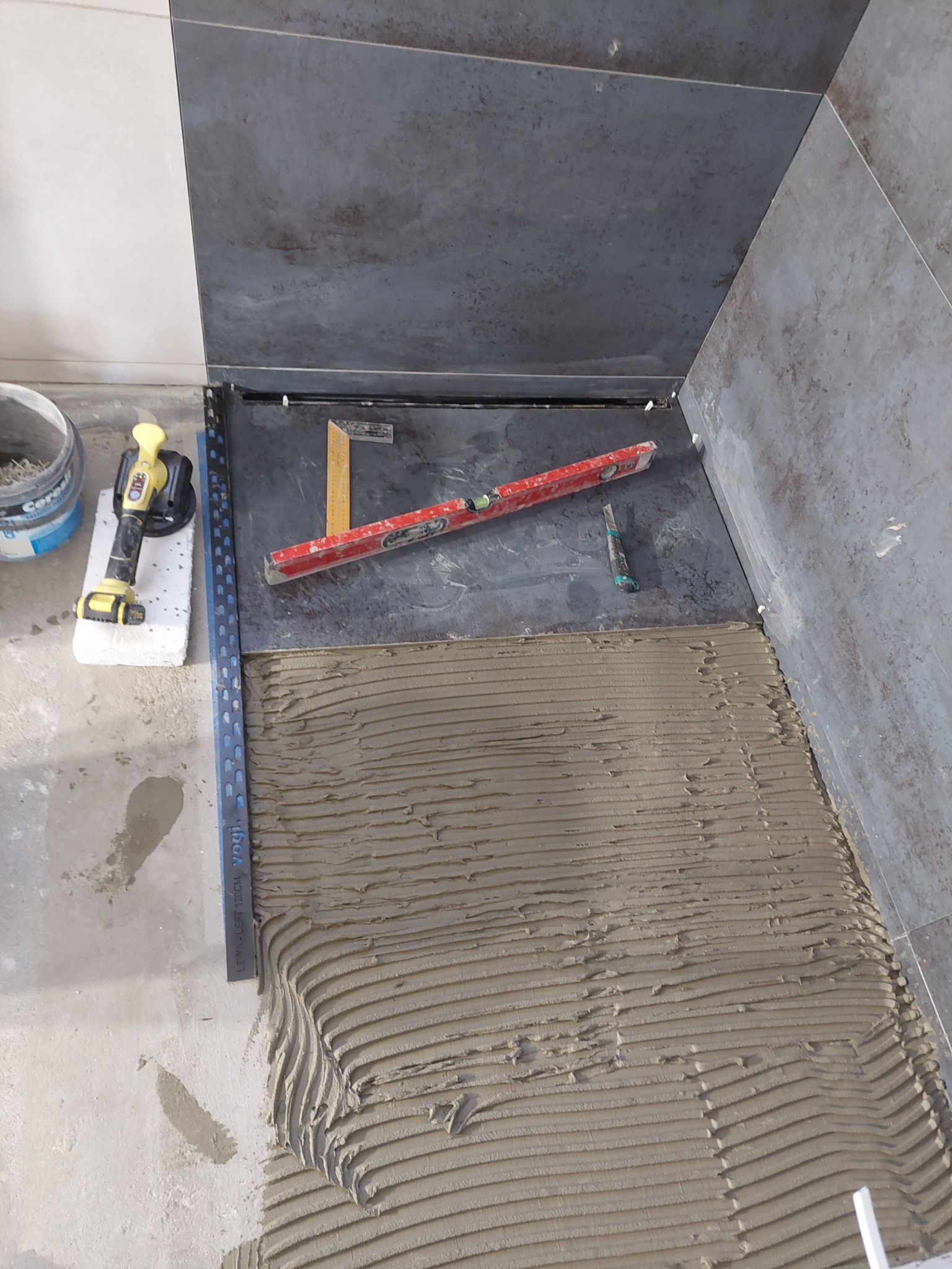 Przygotowanie podłoża pod ciemne płytki w rogu łazienki, widoczna warstwa kleju z charakterystycznymi rowkami, poziomica, kątownik i przyssawka do płytek