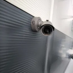 Szara kamera monitoringu zamontowana na szarej, poziomo żłobkowanej ścianie budynku z białym narożnikiem, widok z dołu.