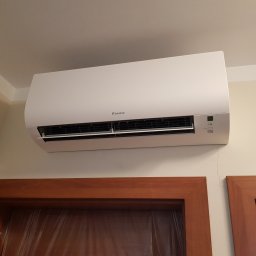 Komi International Michał Kowalski - Biała jednostka klimatyzacji Daikin zamontowana na ścianie nad drewnianą framugą drzwi, widoczna drobna rysa na ścianie.