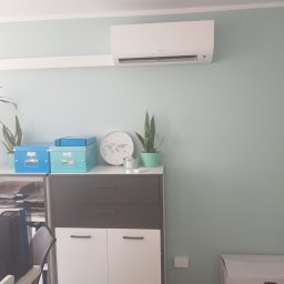 Komi International Michał Kowalski - Biały klimatyzator Daikin zamontowany na ścianie w pokoju biurowym o pastelowych ścianach, nad szafką z dekoracjami i roślinami doniczkowymi.