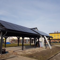 Komi International Michał Kowalski - Montaż paneli fotowoltaicznych na drewnianej wiacie w Opolu, z widoczną drabiną i częściowo ułożonymi panelami, na tle placu zabaw i budynków.