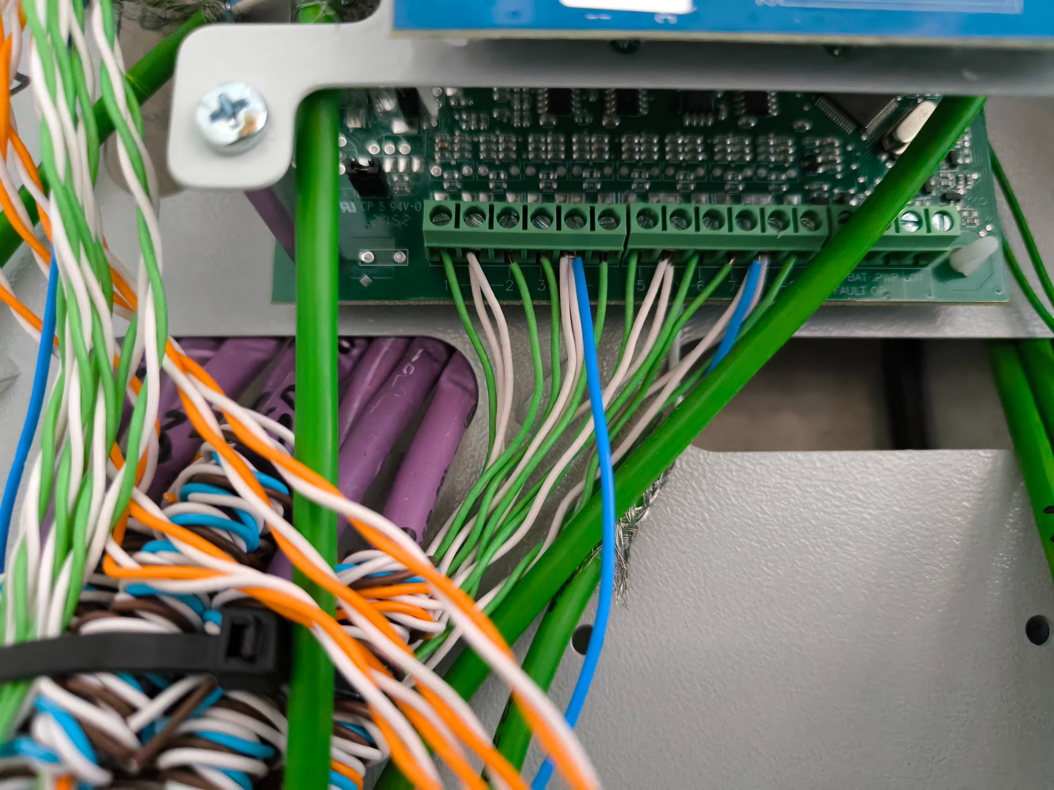 Zbliżenie na panel sterowania z podłączonymi przewodami o różnych kolorach: zielonym, białym, pomarańczowym, niebieskim. Widoczne złącza śrubowe i fragment płytki drukowanej.