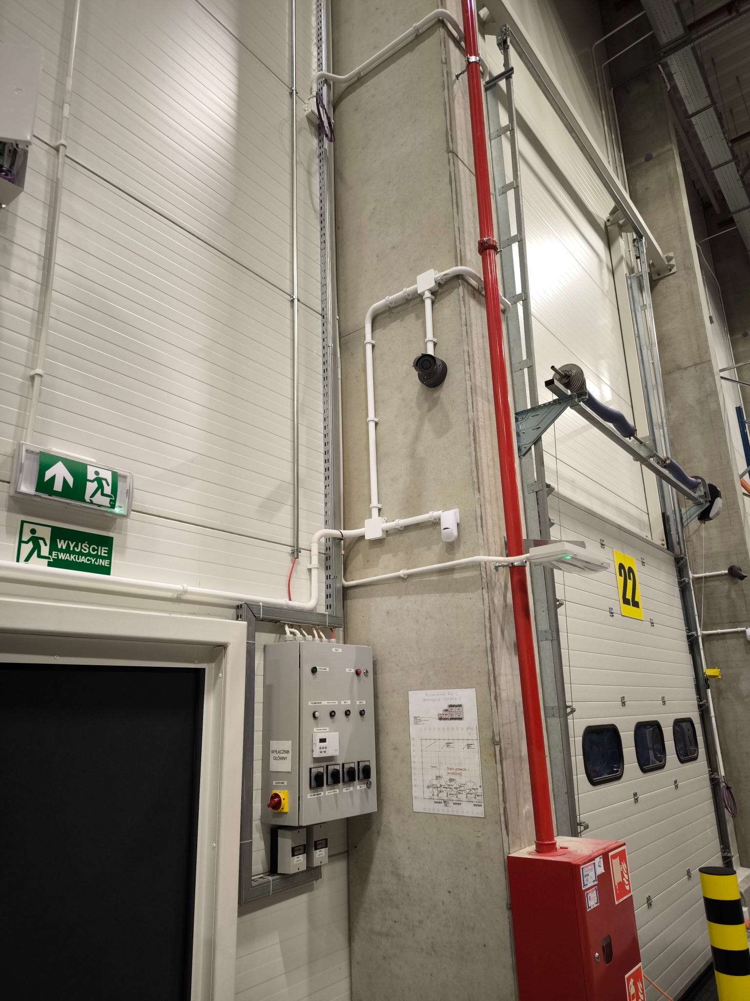 Wnętrze hali magazynowej z widocznym wyjściem ewakuacyjnym, instalacją elektryczną, czujnikiem ruchu, kamerą monitoringu, hydrantem przeciwpożarowym i bramą segmentową z numerem 22.