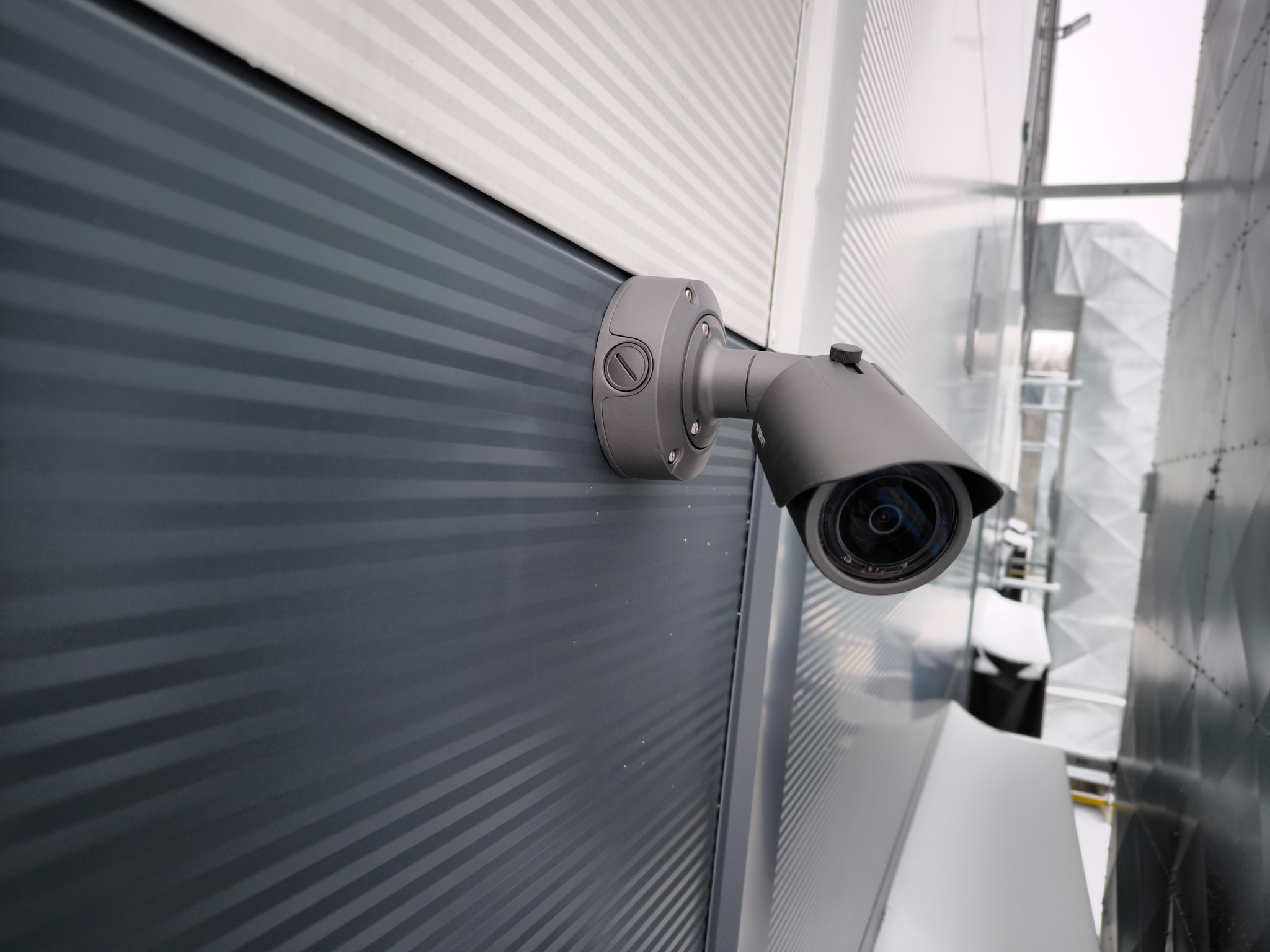Zewnętrzna kamera monitoringu CCTV zamontowana na szarej, metalowej ścianie budynku z widocznymi elementami wentylacji w tle.