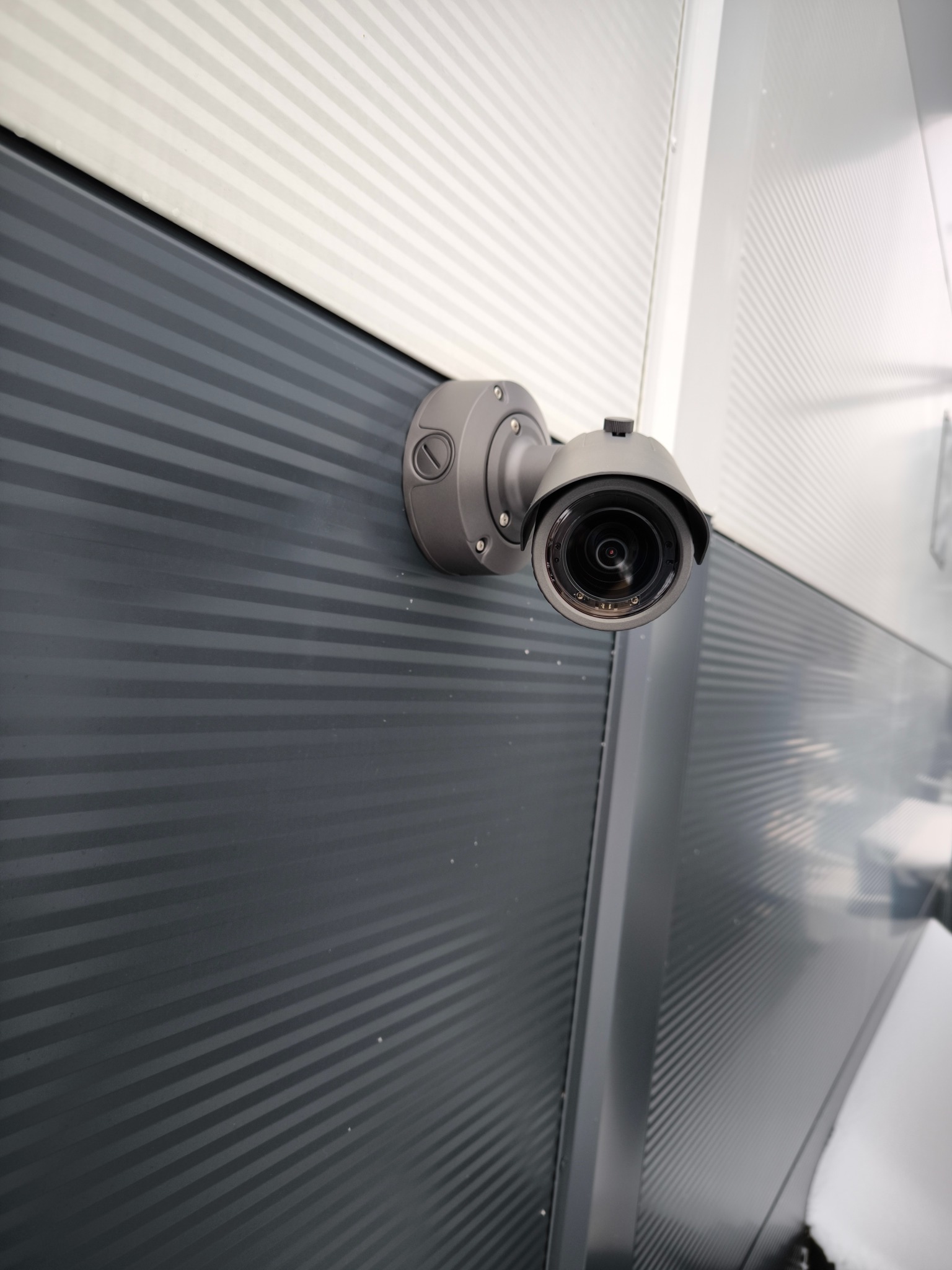 Szara kamera monitoringu zamontowana na szarej, poziomo żłobkowanej ścianie budynku z białym narożnikiem, widok z dołu.