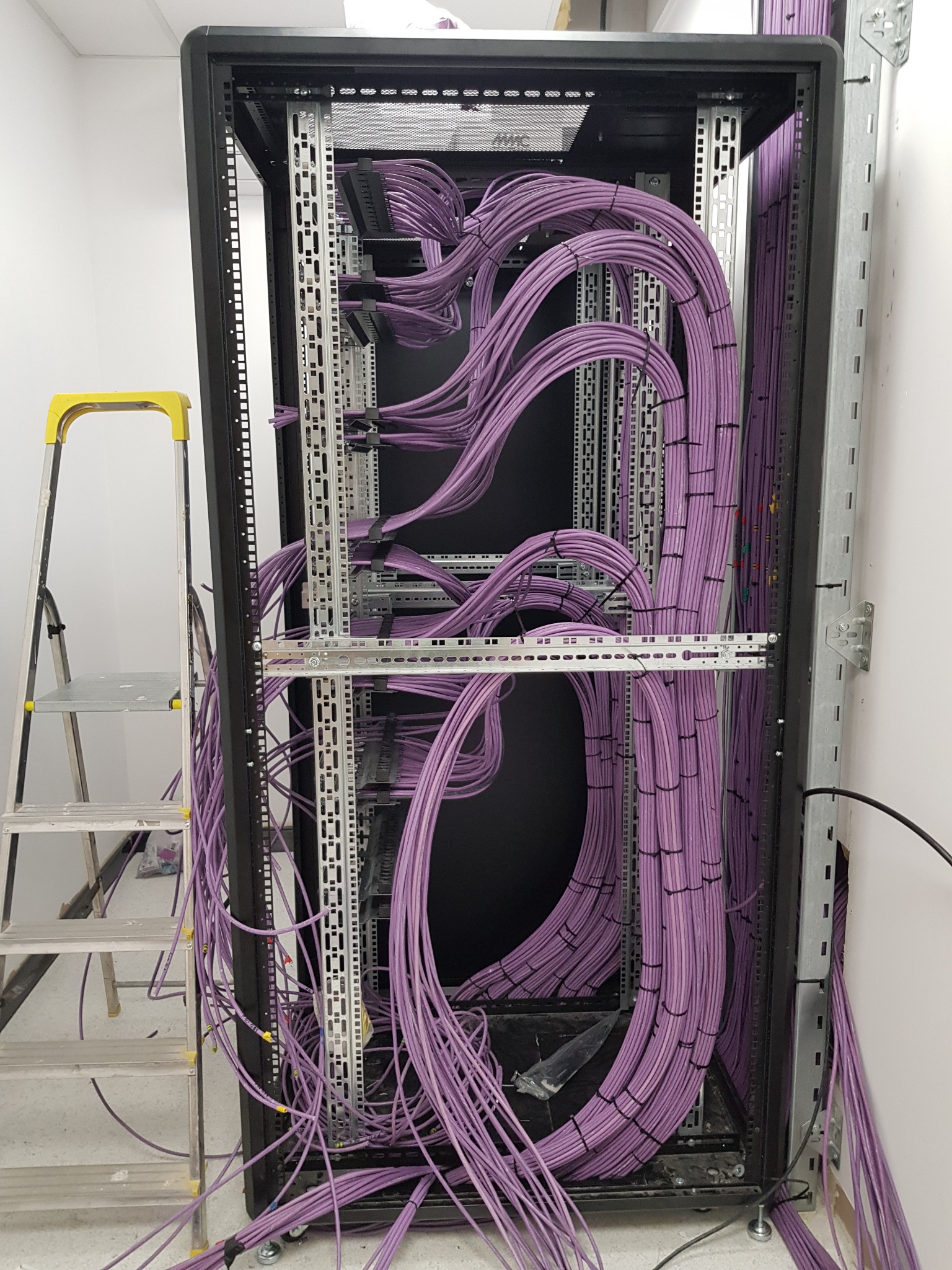 Serwerownia w trakcie instalacji okablowania strukturalnego; fioletowe kable uporządkowane wewnątrz szafy rack, drabina aluminiowa obok.