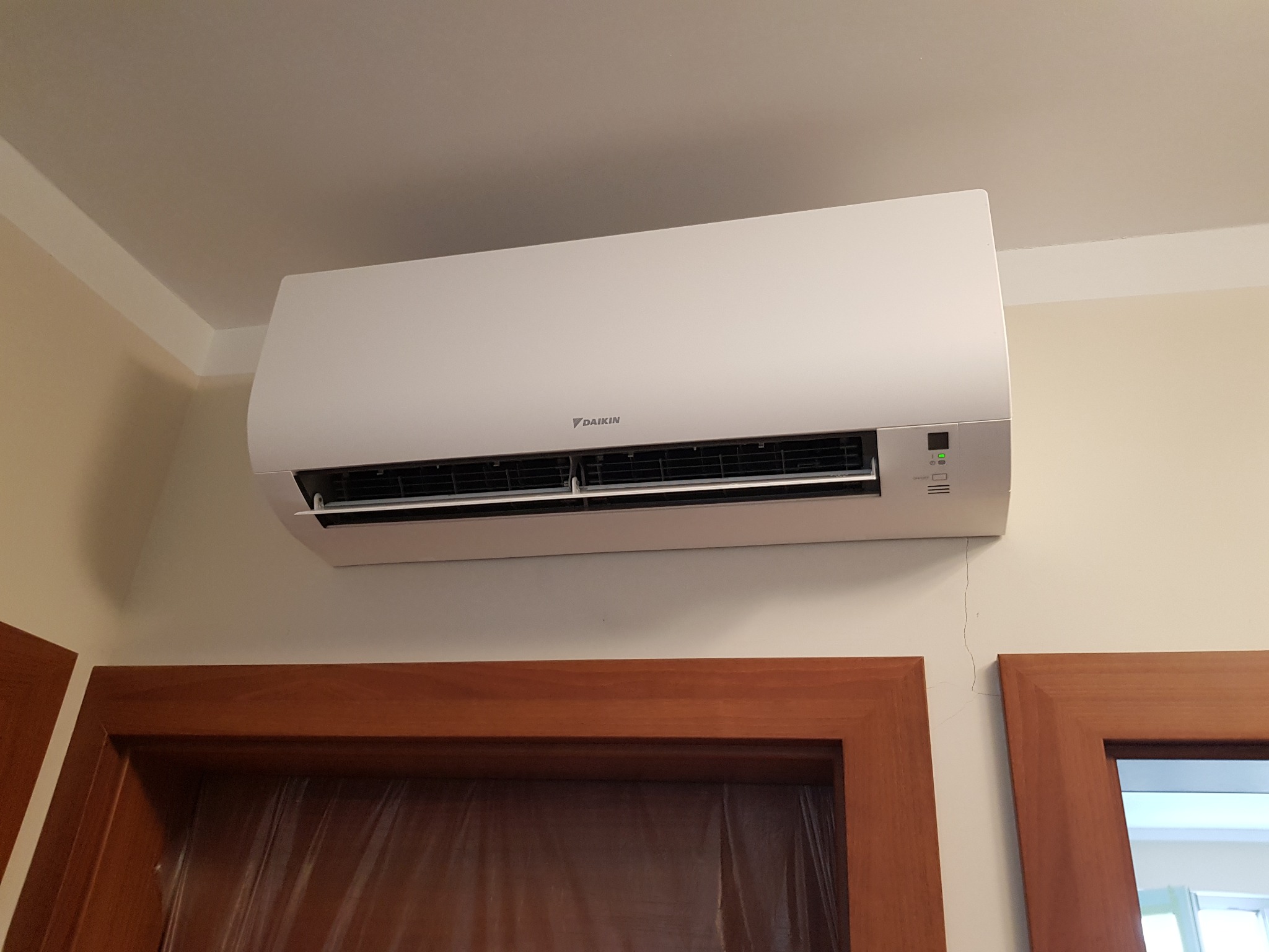 Biała jednostka klimatyzacji Daikin zamontowana na ścianie nad drewnianą framugą drzwi, widoczna drobna rysa na ścianie.