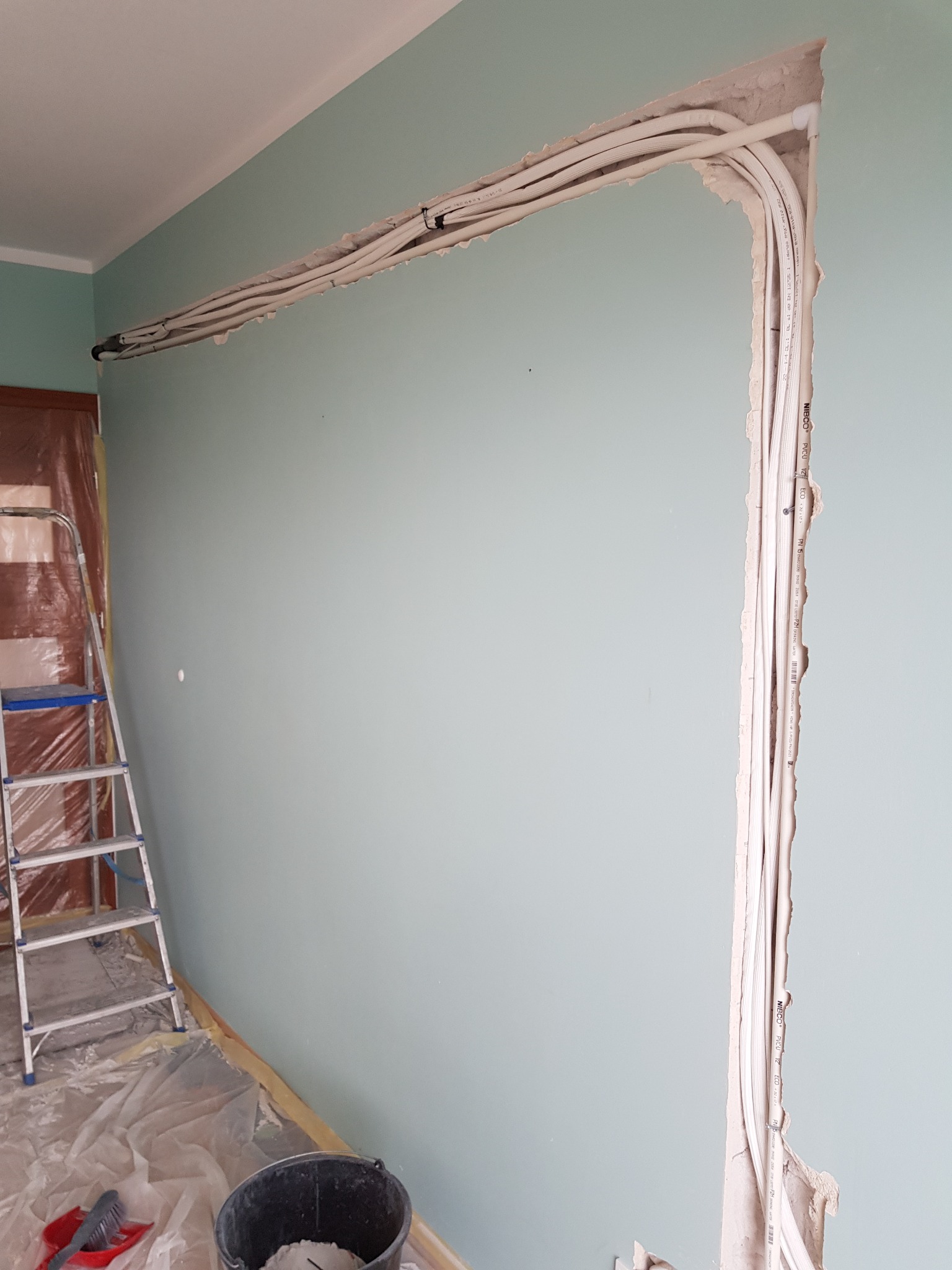 Wykute bruzdy w ścianie w kolorze miętowym, w których ułożono wiązki białych kabli elektrycznych, widoczna drabina i wiadro z zaprawą, przygotowanie do tynkowania.