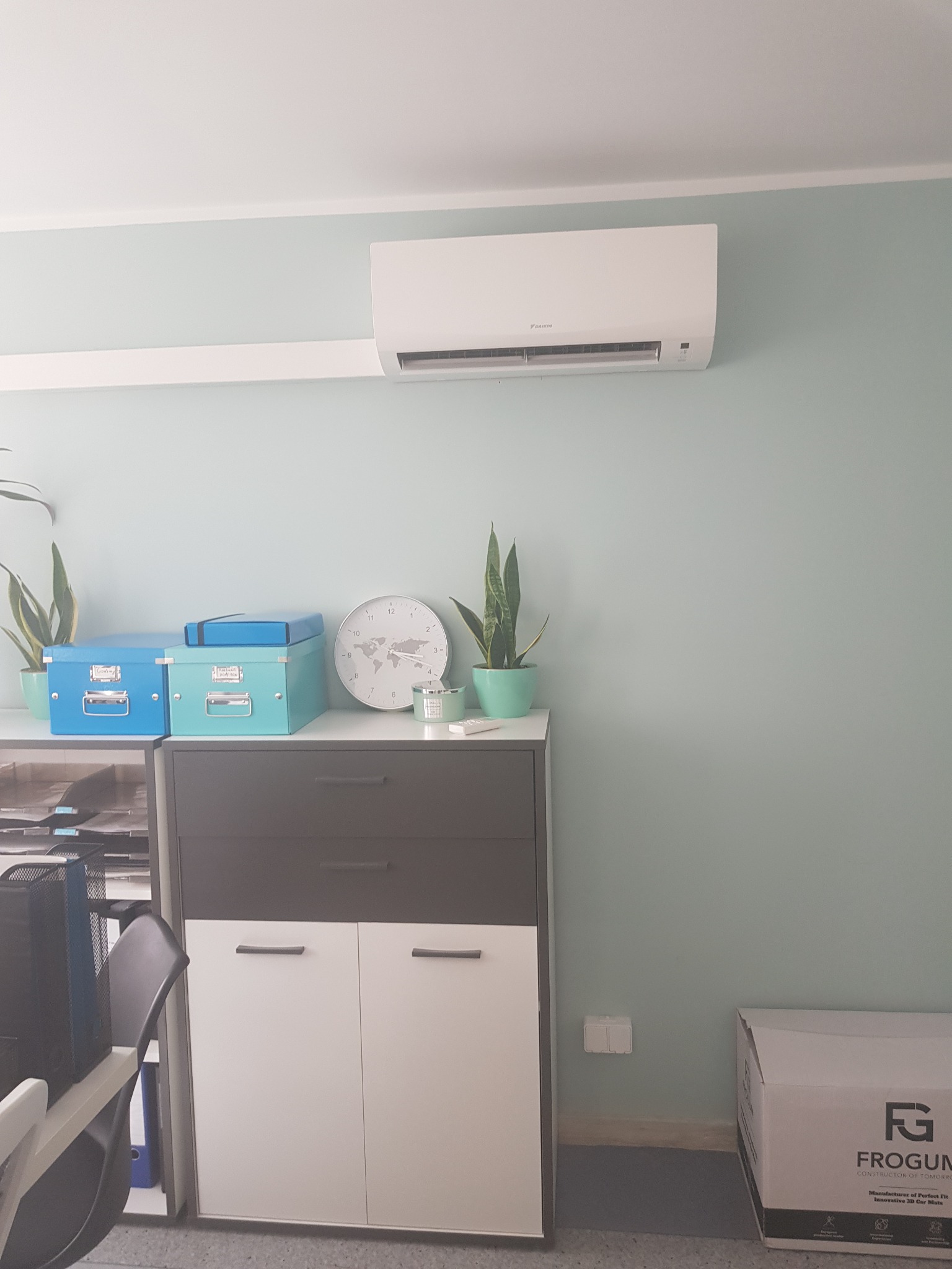 Biały klimatyzator Daikin zamontowany na ścianie w pokoju biurowym o pastelowych ścianach, nad szafką z dekoracjami i roślinami doniczkowymi.