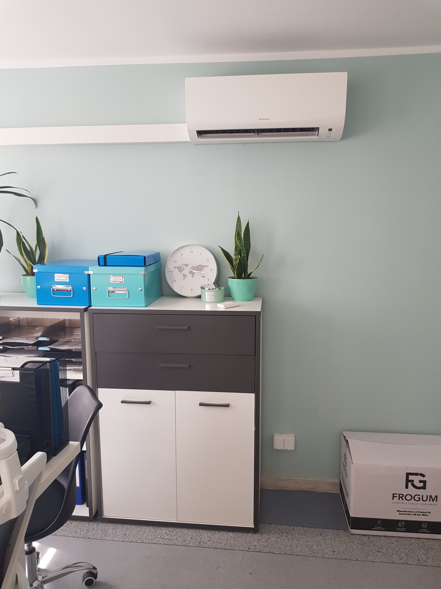 Biały klimatyzator Daikin zamontowany na ścianie w biurze, pod nim szafka z dekoracjami i pudełko Frogum na podłodze.