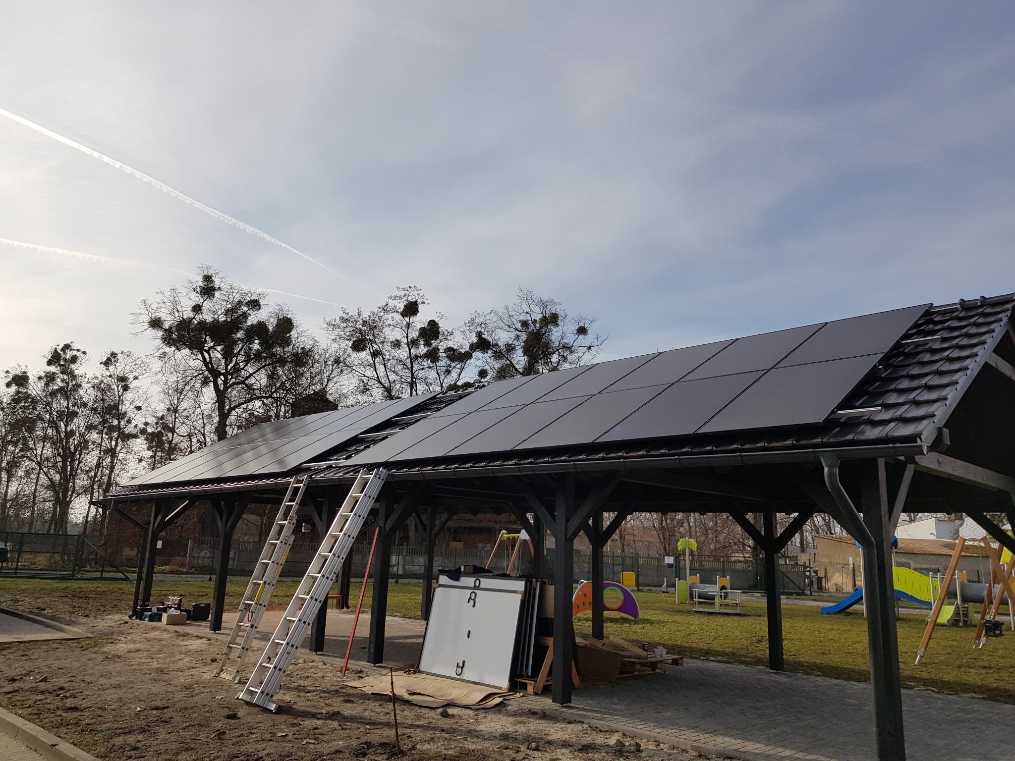 Montaż paneli fotowoltaicznych na drewnianej altanie w parku, widoczne drabiny i panele częściowo zainstalowane, plac zabaw w tle.