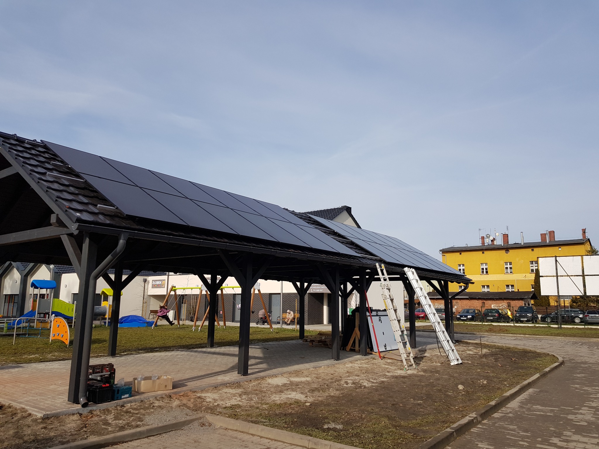 Montaż paneli fotowoltaicznych na drewnianej wiacie w Opolu, z widoczną drabiną i częściowo ułożonymi panelami, na tle placu zabaw i budynków.
