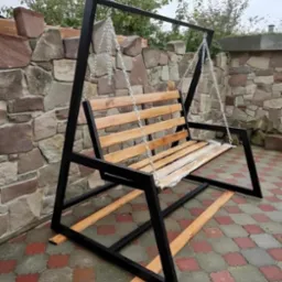 Metalowa huśtawka ogrodowa z drewnianym siedziskiem i oparciem, zawieszona na łańcuchach, z widocznym zabezpieczeniem folią na siedzisku, na tle kamiennej ściany i kostki brukowej.