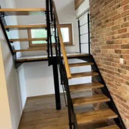 Drewniane schody z czarną metalową konstrukcją, zamocowane do ceglanej ściany, z drewnianą poręczą i widocznym oknem w tle.