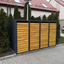 Nowoczesna zabudowa śmietnikowa z metalową ramą i drewnianymi panelami, otoczona zielenią, na tle kostki brukowej i budynku mieszkalnego.