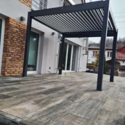 Nowoczesna, czarna pergola z regulowanymi lamelami na tarasie wyłożonym płytami imitującymi drewno, przylegająca do domu z ceglanym akcentem na elewacji.