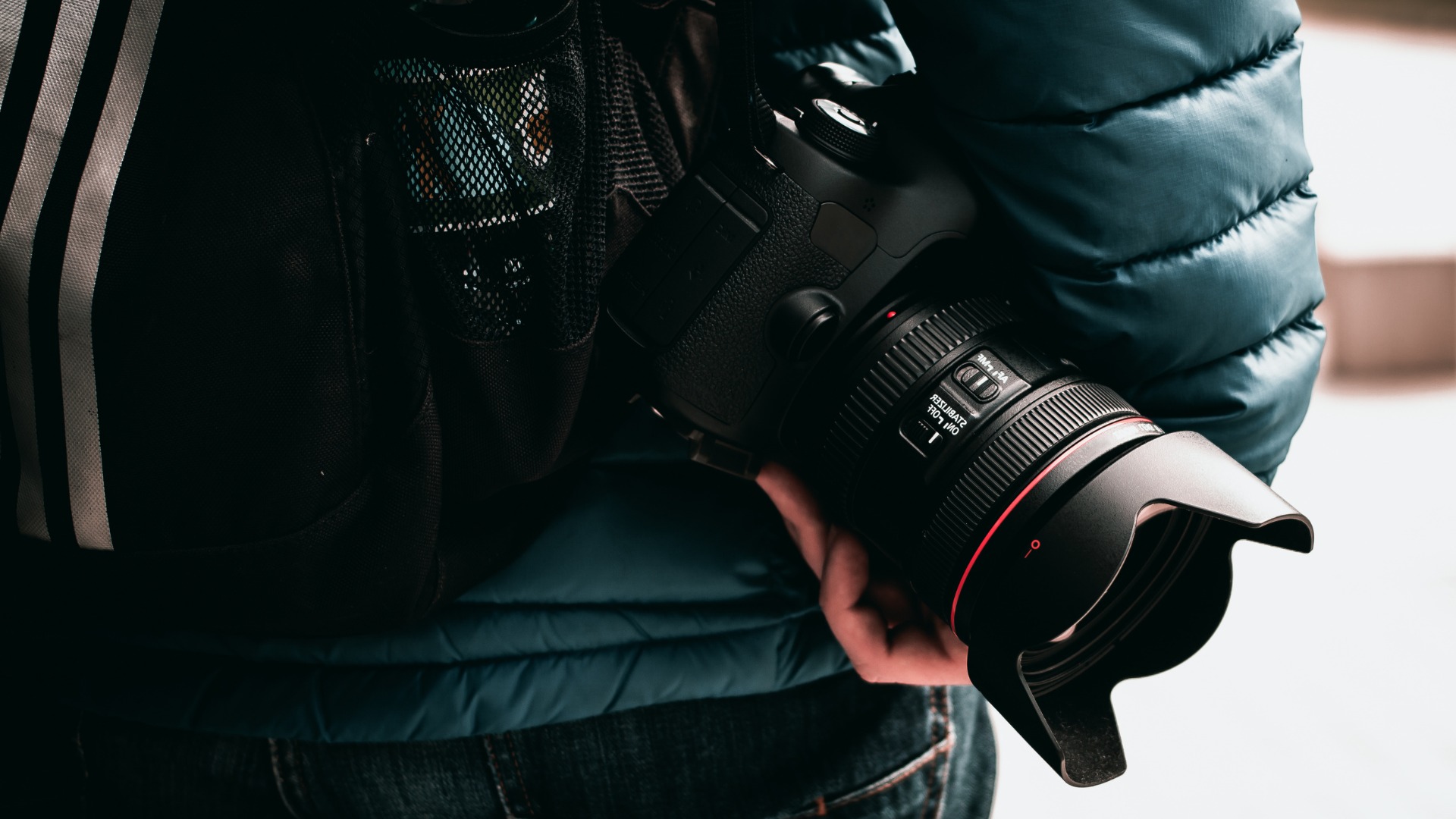 Czarny aparat z obiektywem, osłoną przeciwsłoneczną i czerwonym akcentem, trzymany przez osobę w jeansach i niebieskiej kurtce puchowej, widok zza pleców z plecakiem.