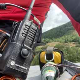 Widok z góry na dach, z radiem Motorola DP1400 w skórzanym futerale, butelką wody Exaler zawieszoną na linie oraz elementami uprzęży a w tle zalesione wzgórze i pochmurne niebo.