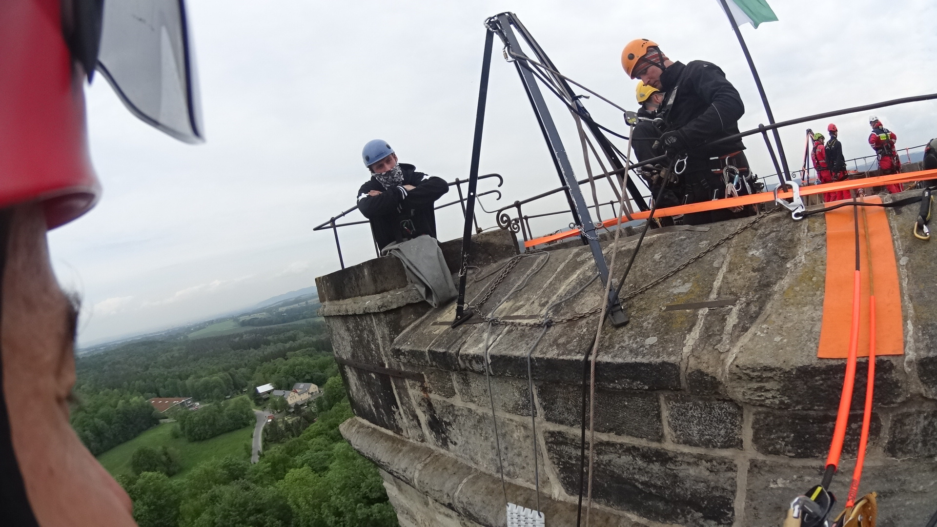 Ekipa alpinistyczna zabezpiecza zabytkową wieżę zamkową, widok z góry na krajobraz zalesionych wzgórz w tle.