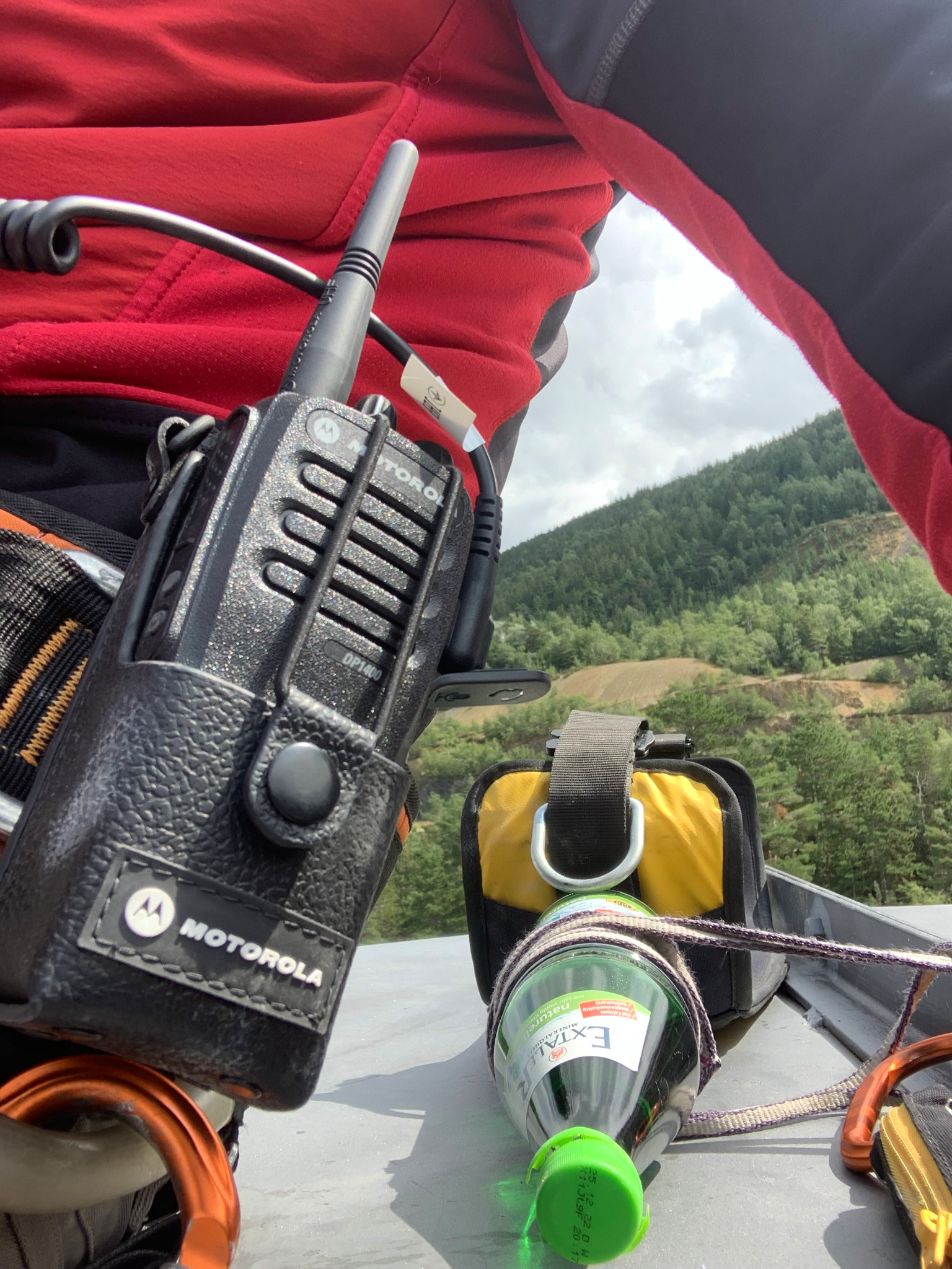 Widok z góry na dach, z radiem Motorola DP1400 w skórzanym futerale, butelką wody Exaler zawieszoną na linie oraz elementami uprzęży a w tle zalesione wzgórze i pochmurne niebo.