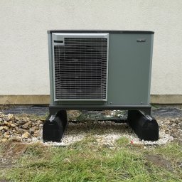 TERM-HOUSE WOJCIECH NOWAK - Zewnętrzna jednostka pompy ciepła Nibe F2040, umieszczona na czarnych podkładkach na żwirowym podłożu, z fragmentem trawnika i elewacji budynku w tle.